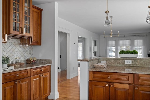 kitchen with hanging light fixtures, glass insert cabinets, brown cabinetry, and light stone counters