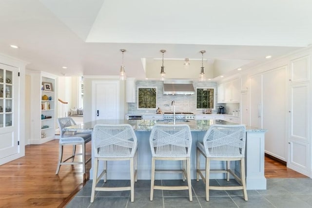 kitchen featuring a center island with sink, white cabinets, wall chimney exhaust hood, light stone counters, and decorative light fixtures