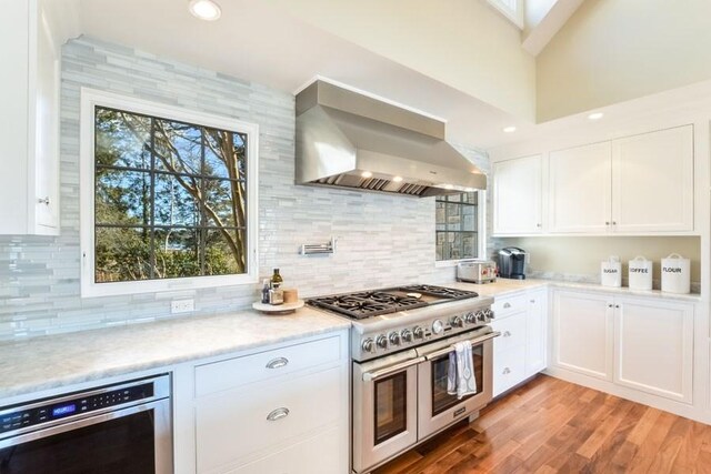 kitchen with wall chimney exhaust hood, light wood-style floors, white cabinetry, and double oven range