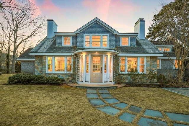 back of house at dusk featuring stone siding, a chimney, and a lawn