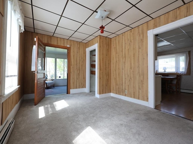 carpeted empty room with a baseboard radiator, a drop ceiling, and wooden walls