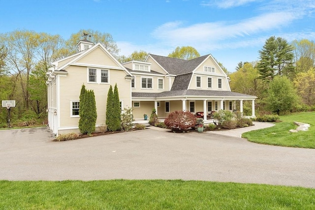 shingle-style home featuring covered porch, a front yard, a chimney, a shingled roof, and asphalt driveway