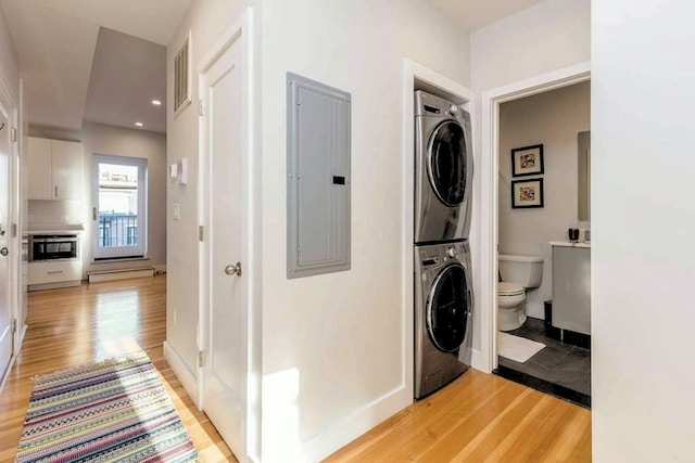 laundry room featuring baseboards, light wood-type flooring, electric panel, laundry area, and stacked washing maching and dryer