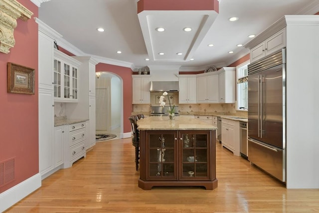 kitchen featuring arched walkways, crown molding, white cabinetry, stainless steel built in refrigerator, and exhaust hood