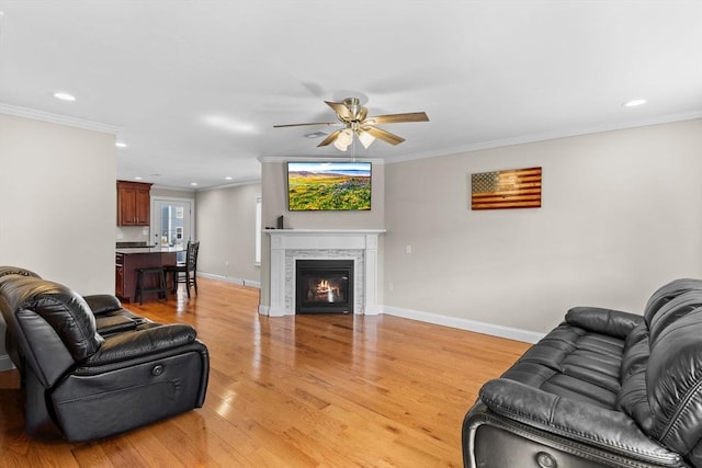 living room with light wood-type flooring, a ceiling fan, a glass covered fireplace, crown molding, and baseboards