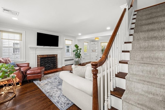 living area with visible vents, crown molding, a brick fireplace, dark wood finished floors, and stairs
