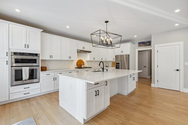 kitchen with appliances with stainless steel finishes, a kitchen island with sink, white cabinetry, light hardwood / wood-style floors, and decorative light fixtures