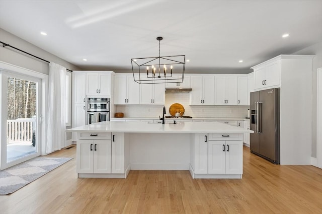 kitchen with appliances with stainless steel finishes, white cabinetry, an island with sink, hanging light fixtures, and light wood-type flooring