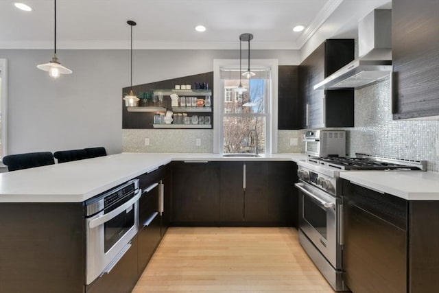 kitchen with light wood-style floors, a peninsula, stainless steel appliances, wall chimney range hood, and a sink