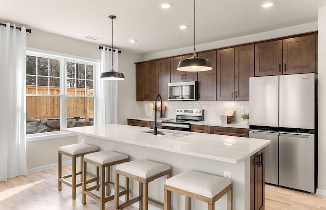 kitchen with light stone counters, dark brown cabinets, hanging light fixtures, stainless steel appliances, and a kitchen island with sink