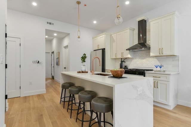 kitchen with stove, wall chimney range hood, pendant lighting, light hardwood / wood-style floors, and an island with sink