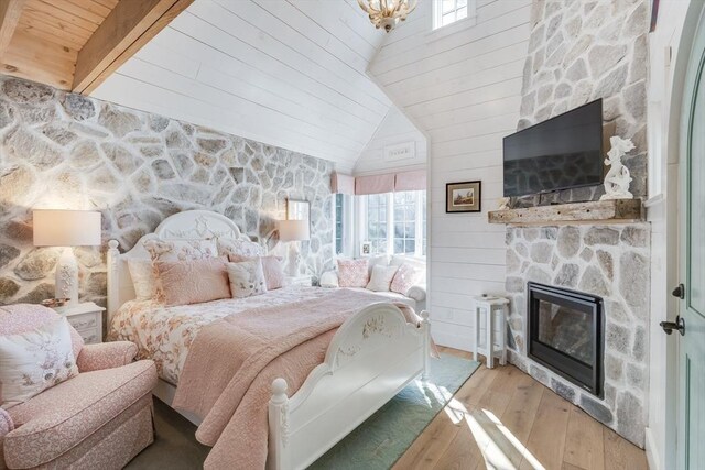 bedroom with multiple windows, a stone fireplace, vaulted ceiling with beams, and light wood-type flooring