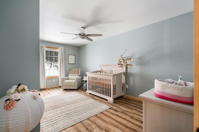 bedroom featuring a nursery area, a ceiling fan, baseboards, and wood finished floors