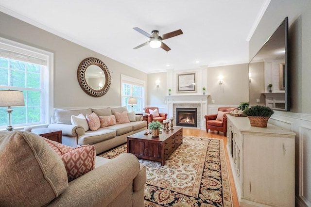 living area featuring a ceiling fan, a decorative wall, a wainscoted wall, light wood finished floors, and a glass covered fireplace