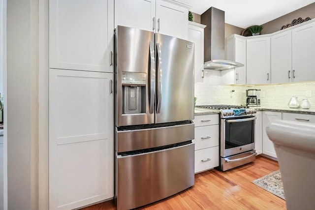 kitchen with appliances with stainless steel finishes, wall chimney exhaust hood, white cabinetry, light wood-style flooring, and tasteful backsplash