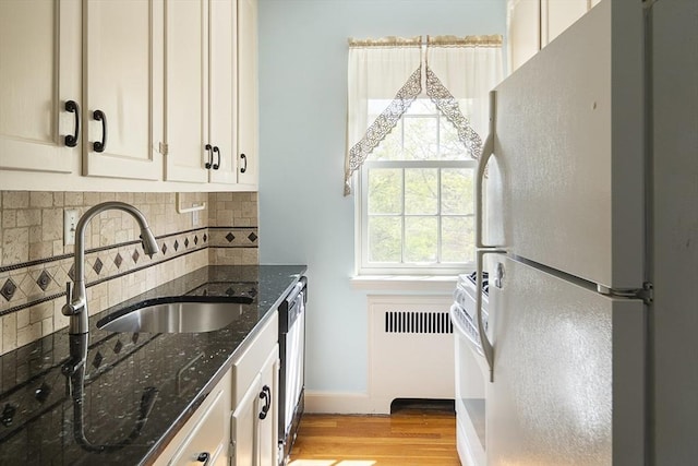 kitchen with a sink, white appliances, dark stone counters, light wood finished floors, and backsplash