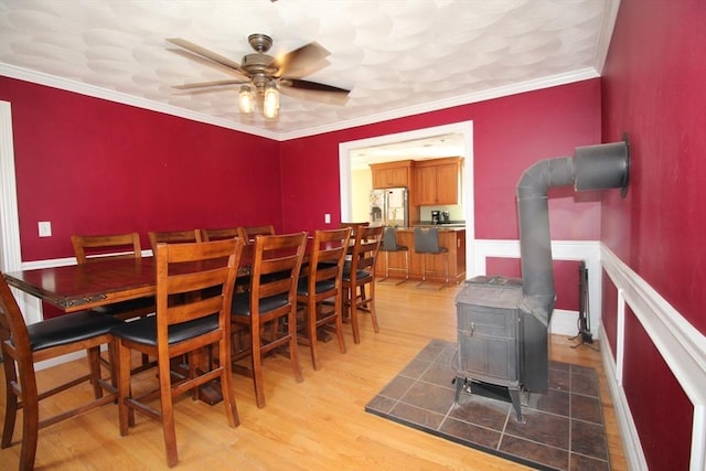 dining space featuring light wood finished floors, a wainscoted wall, ornamental molding, a wood stove, and a ceiling fan