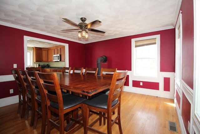 dining space featuring visible vents, light wood-style flooring, ornamental molding, wainscoting, and ceiling fan