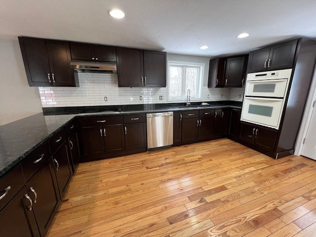 kitchen featuring dishwasher, light wood-style flooring, dark stone countertops, under cabinet range hood, and a sink