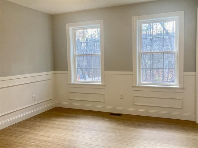 empty room featuring visible vents, light wood-style flooring, a wainscoted wall, and a healthy amount of sunlight