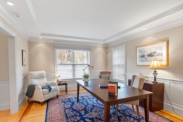 home office featuring light wood-style floors, a wainscoted wall, a decorative wall, crown molding, and a tray ceiling