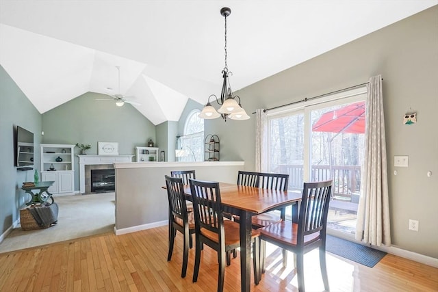 dining room with a tiled fireplace, light wood-style flooring, ceiling fan with notable chandelier, baseboards, and vaulted ceiling