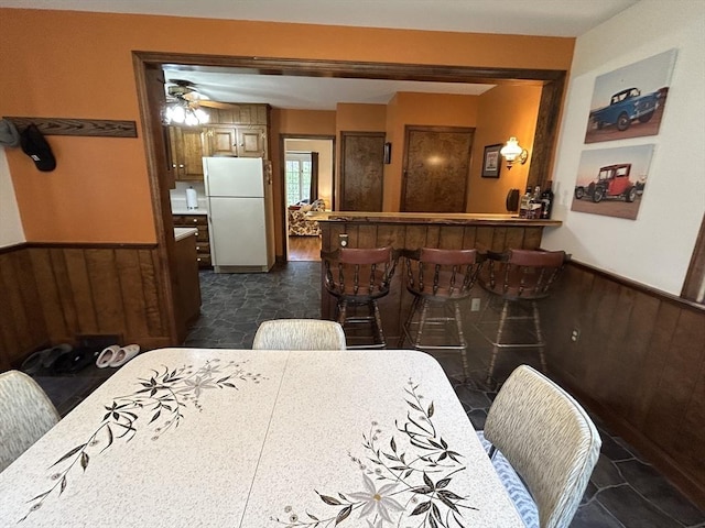 dining space with wooden walls, ceiling fan, wainscoting, stone finish floor, and a bar