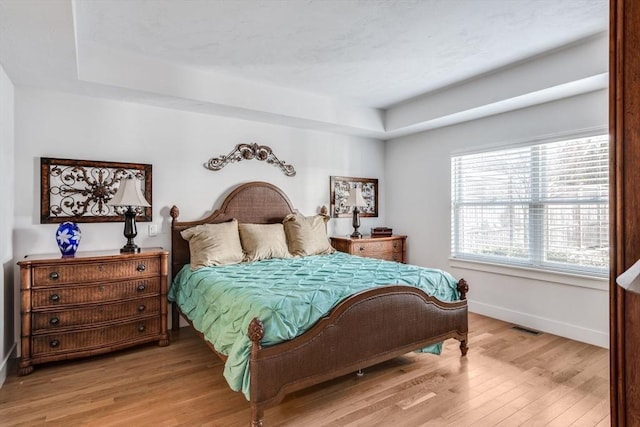 bedroom featuring light wood-type flooring, a raised ceiling, visible vents, and baseboards