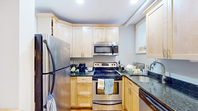 kitchen with a sink, dark stone counters, appliances with stainless steel finishes, and light brown cabinetry