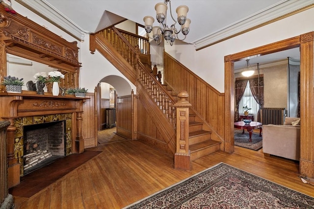 stairs with arched walkways, a wainscoted wall, wood finished floors, a tiled fireplace, and crown molding