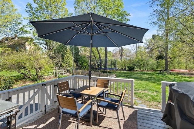 wooden terrace with a lawn, outdoor dining area, a trampoline, and fence