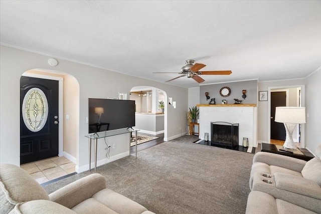 carpeted living area with ceiling fan, a fireplace, arched walkways, baseboards, and ornamental molding