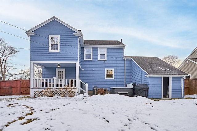 snow covered back of property with roof with shingles, a jacuzzi, and fence