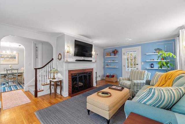 living area featuring arched walkways, light wood-style floors, stairway, a brick fireplace, and crown molding