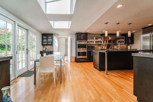 kitchen with appliances with stainless steel finishes, light wood-style floors, wall chimney exhaust hood, backsplash, and a skylight