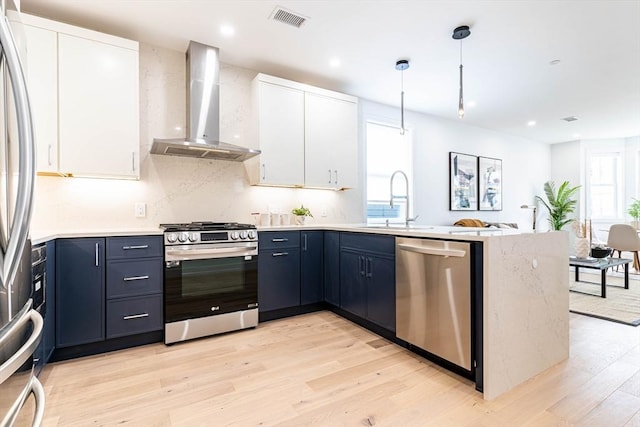 kitchen with stainless steel appliances, blue cabinets, wall chimney range hood, decorative light fixtures, and white cabinets