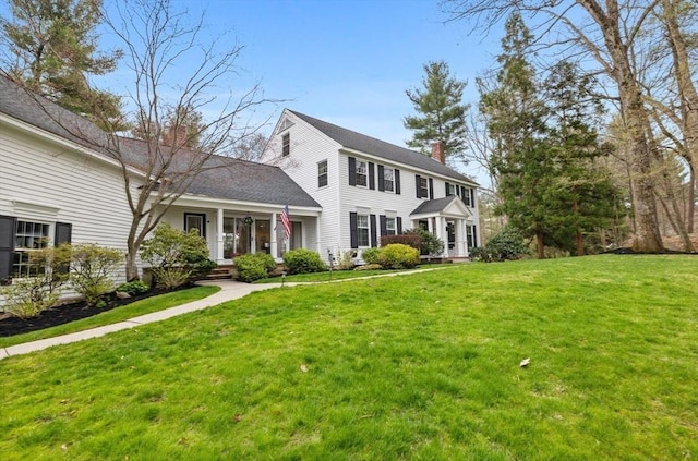 colonial-style house with a shingled roof, a front yard, a chimney, and covered porch