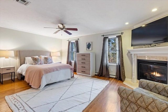 bedroom with ceiling fan, a baseboard heating unit, light hardwood / wood-style floors, a tiled fireplace, and ornamental molding