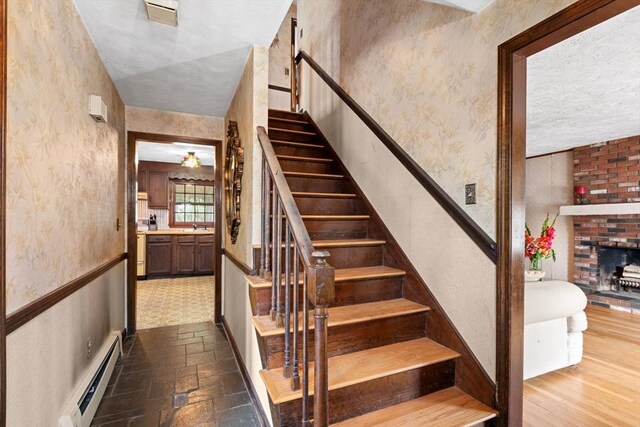 stairway featuring a baseboard heating unit, wallpapered walls, and a brick fireplace