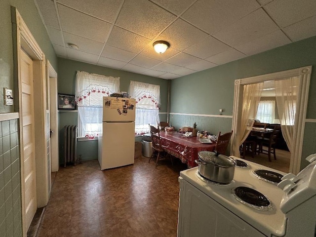 kitchen featuring white appliances, radiator heating unit, a wealth of natural light, and a drop ceiling