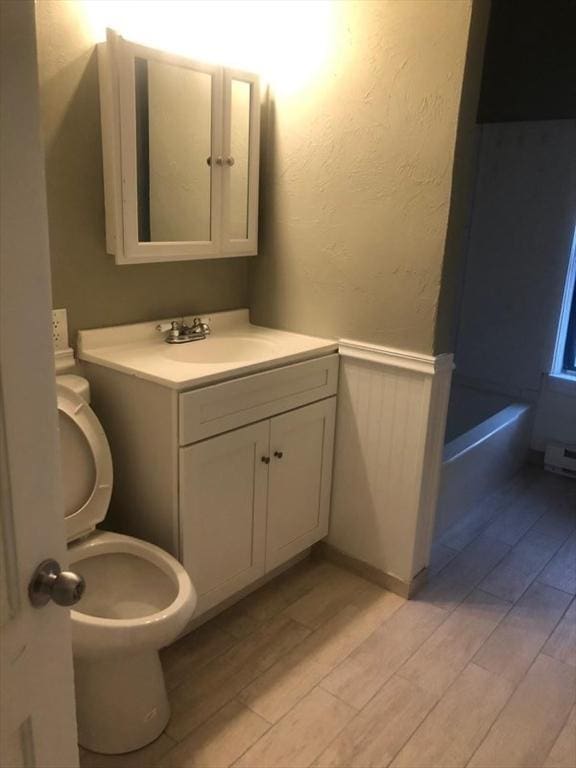 bathroom with a textured wall, toilet, wood finished floors, vanity, and wainscoting