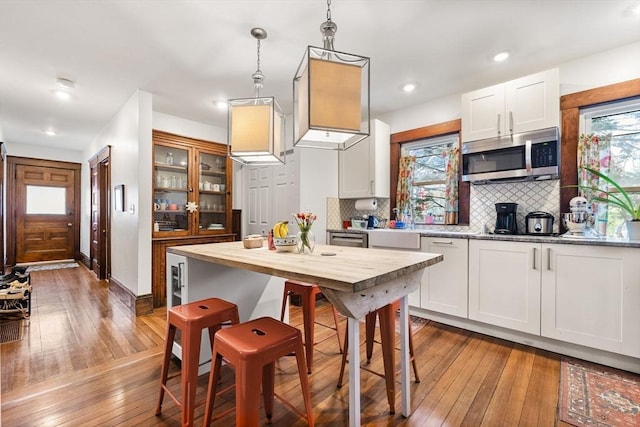 kitchen featuring wood-type flooring, decorative backsplash, stainless steel microwave, white cabinets, and decorative light fixtures