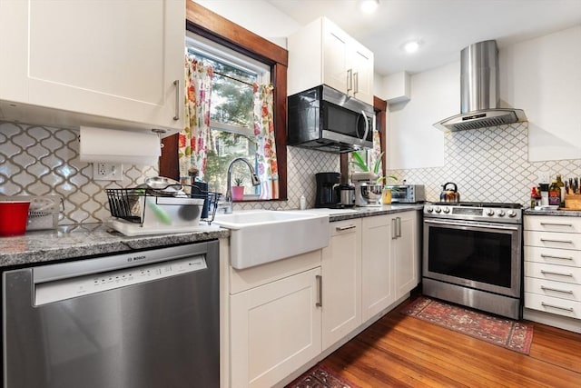 kitchen featuring wall chimney range hood, backsplash, appliances with stainless steel finishes, a sink, and white cabinets
