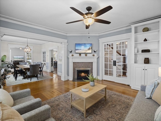 living area with ceiling fan with notable chandelier, baseboards, a fireplace with flush hearth, wood finished floors, and ornamental molding