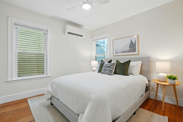 bedroom with a wall unit AC, baseboards, wood finished floors, and ceiling fan