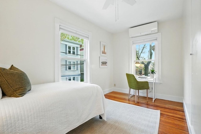 bedroom with a wall mounted AC, wood finished floors, multiple windows, and baseboards