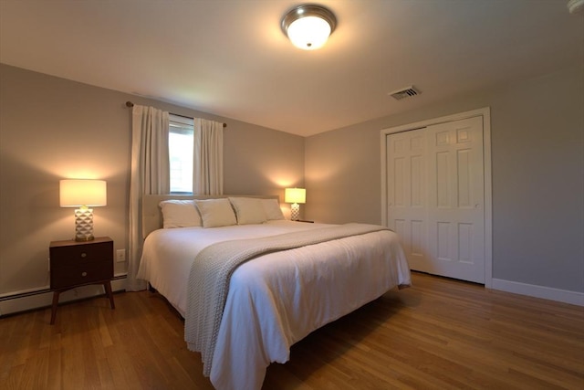bedroom featuring visible vents, a closet, wood finished floors, baseboards, and a baseboard heating unit