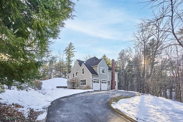 snow covered property with an attached garage, a chimney, and aphalt driveway