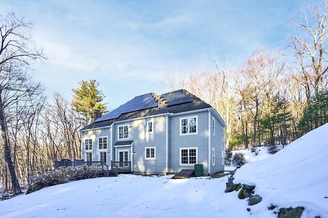 snow covered back of property with a deck, a chimney, and roof mounted solar panels