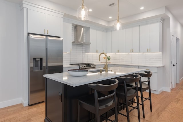 kitchen featuring light hardwood / wood-style flooring, wall chimney exhaust hood, an island with sink, appliances with stainless steel finishes, and light stone counters